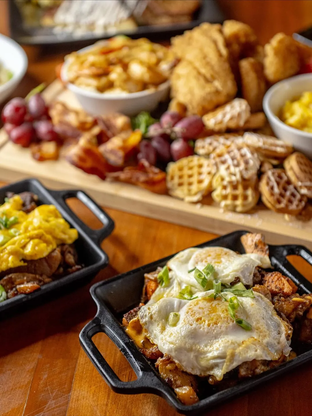 Brunch board of waffles, bacon and fruit beside a skillet topped with a fried egg